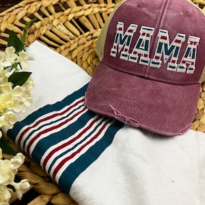 May include: A red and beige baseball cap with a white mesh back. The cap has the word "MAMA" embroidered on the front in white, red, and blue stripes.
