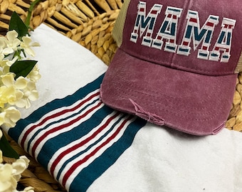 Puede incluir: Una gorra de béisbol granate desgastada con parte trasera de malla y la palabra MAMA bordada en letras de bloque blancas, rojas y verde azulado. La gorra está sobre una tela blanca con rayas horizontales rojas y verde azulado y flores blancas.