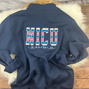 May include: Navy blue quarter-zip sweatshirt with the word "NICU" in a block letter design, featuring red and blue stripes. Below the word "NICU" is the word "nurse" in cursive with two hearts on either side. The sweatshirt is laying on a white furry surface.