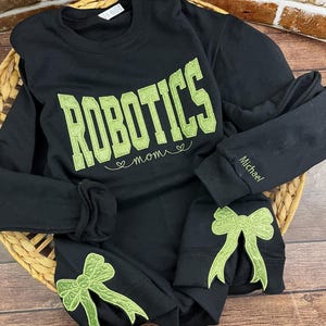 May include: Black sweatshirt with green embroidered text that reads "Robotics Mom" and green embroidered bows on the sleeves. The text "Michael" is embroidered on one sleeve.