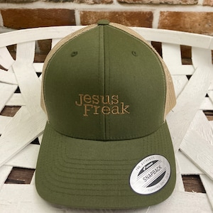 May include: A green and tan baseball cap with the text "Jesus Freak" embroidered on the front. The cap has a mesh back and a "The Authentic Yupoong Snapback" label on the back.