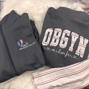 May include: A dark gray zip-up jacket featuring embroidered blue and pink baby footprints with the text Kim S. BSN, RN, RNC-OB, C-EFM, alongside a dark gray sweatshirt with the text OBGYN midwife and a white fabric with pink and gray stripes.