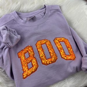 May include: A light purple crewneck sweatshirt featuring the word BOO in large, block-style letters across the chest. The letters are filled with bright orange sequins and outlined with a thin orange border, creating a textured, reflective appearance.