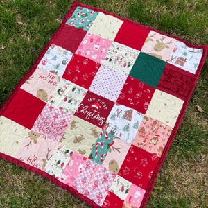 May include: A patchwork Christmas quilt with a red border. The quilt features various festive patterns, including reindeer, gingerbread men, and candy canes. The center square reads "My First Christmas."