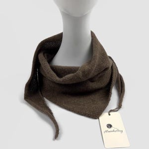 May include: A brown knitted triangular scarf, displayed on a mannequin head. The scarf has a tag that reads "Almaknitting". The scarf is a solid colour and appears to be made of a soft material.
