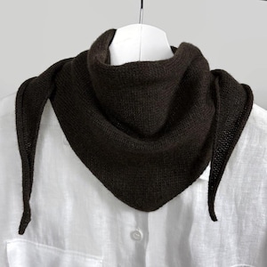 May include: A dark brown triangular scarf, likely knitted, hangs on a white hanger. The scarf has a textured appearance and drapes in a triangular shape, with pointed ends. The scarf is displayed against a white shirt.