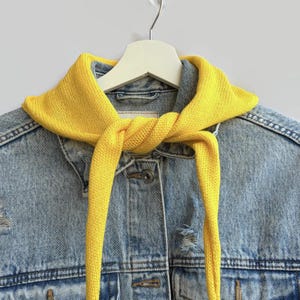 Puede incluir: Un pañuelo triangular de punto amarillo brillante atado con un nudo sencillo alrededor del cuello de una chaqueta vaquera azul claro desgastada. La chaqueta presenta un frente abotonado clásico con botones metálicos y costuras visibles.
