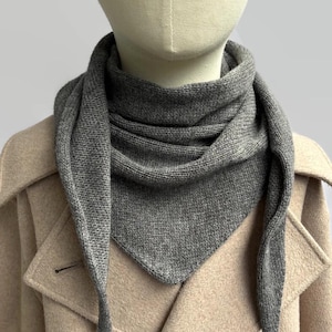 Puede incluir: Una bufanda triangular de punto gris con un patrón tejido texturizado drapeada alrededor del cuello. La bufanda presenta un borde frontal puntiagudo y se lleva sobre un abrigo de color beige claro con solapas anchas y un botón visible.