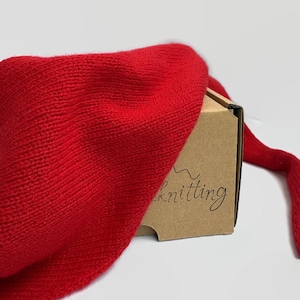 Może przedstawiać: Jasny czerwony wełniany szal położony na kartonowym pudełku z nadrukiem "The Knitting" na boku.