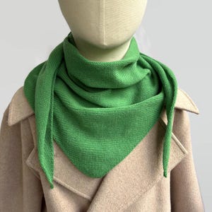 Puede incluir: Una bufanda triangular de punto en color verde brillante drapeada alrededor del cuello de un maniquí. La bufanda presenta un patrón de punto jersey fino y uniforme con un borde limpio, sobre un abrigo de color beige claro con cuello ancho.