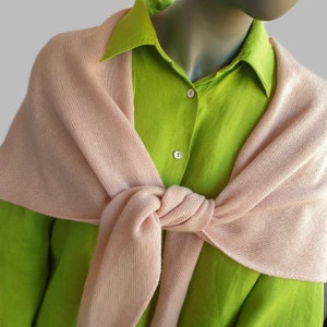 Puede incluir: Una camisa de botones de color verde lima brillante combinada con un suéter de punto rosa pálido suave, colocado sobre los hombros y atado en un nudo en la parte delantera. La camisa tiene un cuello clásico y dos botones blancos visibles.