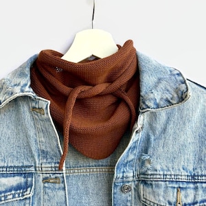 May include: A rust-coloured knitted cowl neck scarf with a drawstring, displayed on a white hanger. The scarf is layered over a light blue denim jacket, creating a contrast in textures and colours. The jacket has silver buttons and a classic design.