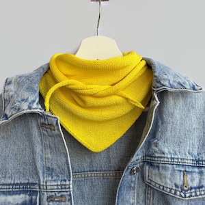 Pode incluir: Um lenço bandana triangular de malha amarelo brilhante sobre uma jaqueta jeans azul claro. O cachecol apresenta uma textura de malha fina e pontas, enquanto a jaqueta jeans exibe uma lavagem clássica clara com botões de metal visíveis.
