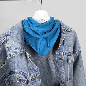 May include: A bright blue knit scarf is draped over a light blue denim jacket. The scarf is triangular and appears to be made of a soft, textured material. The denim jacket has a classic design with button closures and distressed details.