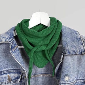 Pode incluir: Um cachecol de malha verde vibrante está pendurado em um cabide branco, contra um fundo cinza claro. O cachecol é triangular, com pontas longas e penduradas. Uma jaqueta jeans azul claro é parcialmente visível, adicionando um contraste casual à textura e cor do cachecol.