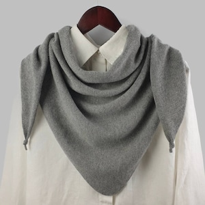 Könnte beinhalten: Ein grauer Strick Schal mit Quasten an den Enden. Der Schal ist über ein weißes Hemd mit Kragen gelegt.