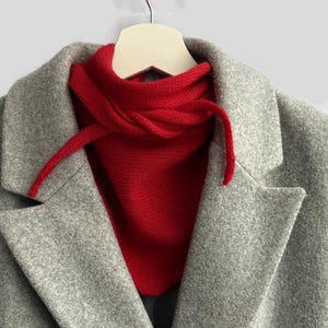 Puede incluir: Un calentador de cuello tejido en color rojo brillante con cordones para atar se lleva debajo de un abrigo de mezcla de lana gris claro. El abrigo presenta solapas anchas y un cuello estructurado, con el accesorio rojo alrededor del cuello.