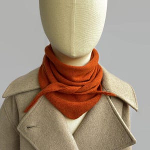Pode incluir: Um lenço triangular de malha em tom laranja queimado vibrante atado ao pescoço de um manequim. O lenço apresenta uma textura de malha fina e consistente, sobreposto a um casaco de mistura de lã bege com lapelas largas e um botão visível.