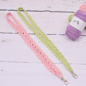 May include: Two crocheted lanyards with silver clasps. One is pink and the other is green. The lanyards are on a white wooden surface with a ball of purple yarn in the background.