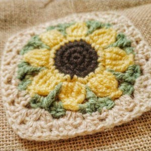 Peut inclure: Un tournesol crocheté avec un centre brun, des pétales jaunes et des feuilles vertes. Le tournesol est entouré d'une bordure beige en crochet.