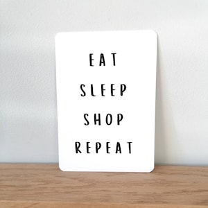 Op de afbeelding: Witte kaart met zwarte tekst die "Eten Slapen Winkelen Herhalen" zegt.