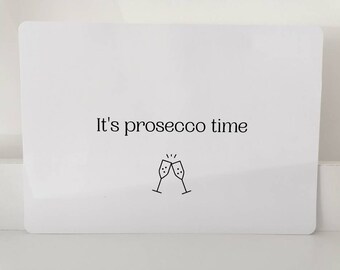 Op de afbeelding: Witte kaart met zwarte tekst die "It's prosecco time" zegt en een zwarte lijntekening van twee champagne glazen die klinken.