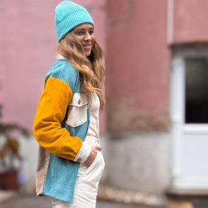 May include: A woman wearing a blue, yellow, and white corduroy jacket, white pants, and a blue beanie stands in front of a pink building. She is smiling and looking at the camera.