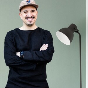 Op de afbeelding: Een man in een zwart shirt met lange mouwen en een bruine baseballpet glimlacht naar de camera. Hij staat voor een groene muur met een zwarte staande lamp.