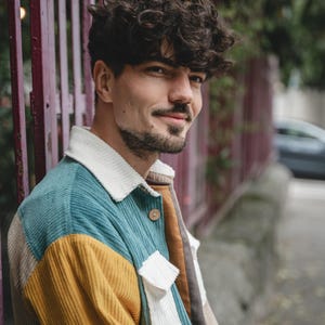 May include: A man with curly brown hair wearing a multi-colored corduroy jacket. The jacket has teal, yellow, white, and brown panels.