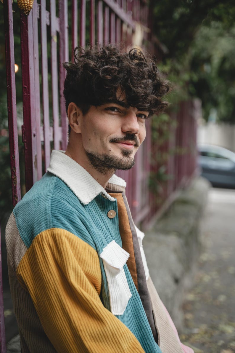May include: A man with curly brown hair wearing a multi-colored corduroy jacket. The jacket has teal, yellow, white, and brown panels.