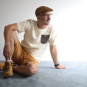 Pu&ograve; includere: Un uomo con un berretto marrone chiaro, una maglietta bianca con una tasca a toppa marrone e pantaloncini marroni &egrave; seduto su un pavimento grigio. Indossa scarpe marroni e un orologio nero.