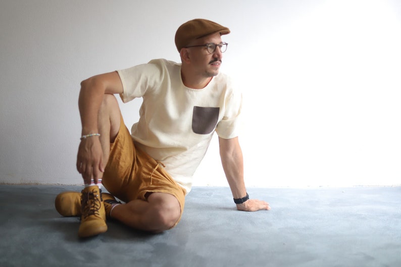 Pu&ograve; includere: Un uomo con un berretto marrone chiaro, una maglietta bianca con una tasca a toppa marrone e pantaloncini marroni &egrave; seduto su un pavimento grigio. Indossa scarpe marroni e un orologio nero.