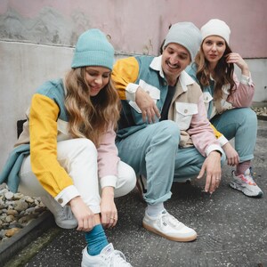 May include: Three people wearing colorful jackets with different color blocks, white pants, and beanies. They are sitting on a sidewalk with a pink wall in the background. Two of the people are wearing white sneakers and one is wearing black and white sneakers.