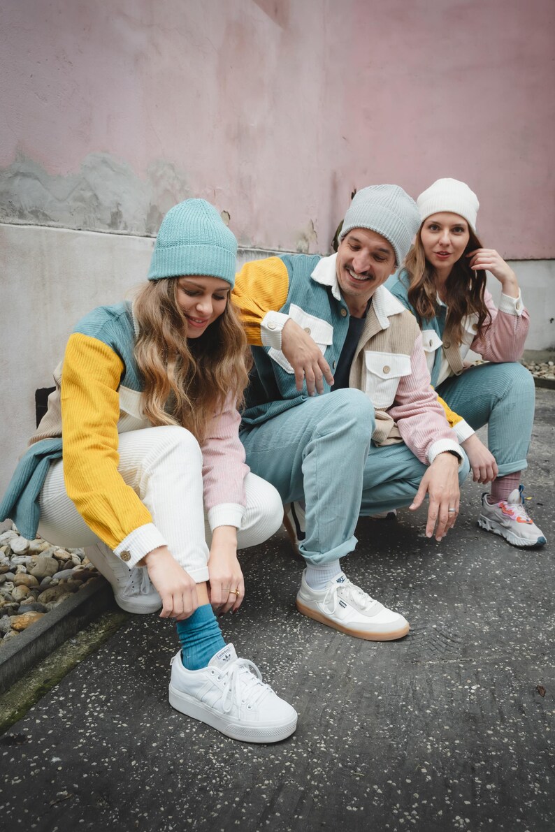 May include: Three people wearing colorful jackets with different color blocks, white pants, and beanies. They are sitting on a sidewalk with a pink wall in the background. Two of the people are wearing white sneakers and one is wearing black and white sneakers.