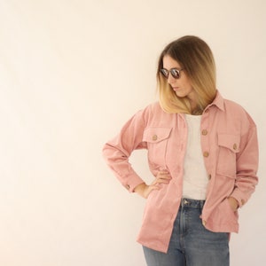 Peut inclure: Une femme portant une veste en velours côtelé rose avec un t-shirt blanc en dessous et un jean bleu. Elle porte des lunettes de soleil et a les mains dans les poches.
