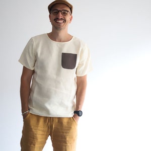 Pu&ograve; includere: Un uomo indossa una camicia di lino bianca con una tasca a toppa marrone, pantaloni di lino giallo senape, un berretto marrone e occhiali.