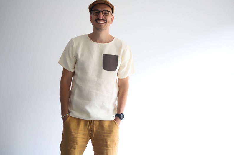 Pu&ograve; includere: Un uomo indossa una camicia di lino bianca con una tasca a toppa marrone, pantaloni di lino giallo senape, un berretto marrone e occhiali.