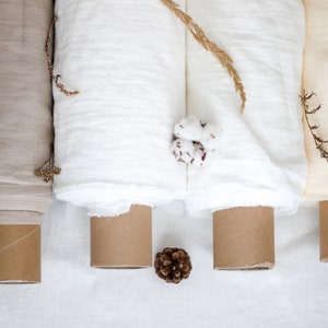 May include: Three rolls of fabric in beige, white, and cream colors are rolled up on cardboard tubes. The fabric is a natural linen material. The rolls are arranged horizontally on a white surface.