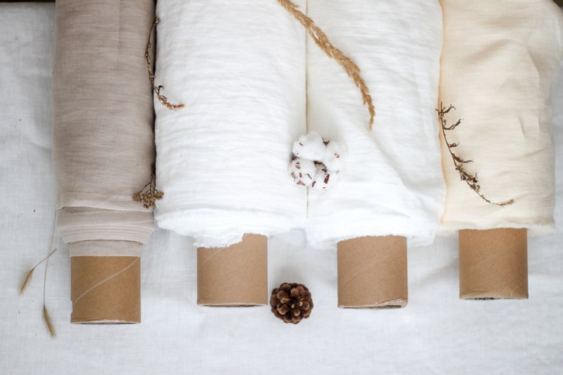 May include: Three rolls of fabric in beige, white, and cream colors are rolled up on cardboard tubes. The fabric is a natural linen material. The rolls are arranged horizontally on a white surface.