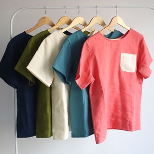 Pu&ograve; includere: Cinque camicie a maniche corte appese a grucce. Le camicie sono di vari colori, tra cui blu navy, verde oliva, crema, blu verde e corallo. Una camicia corallo ha una tasca a toppa bianca.