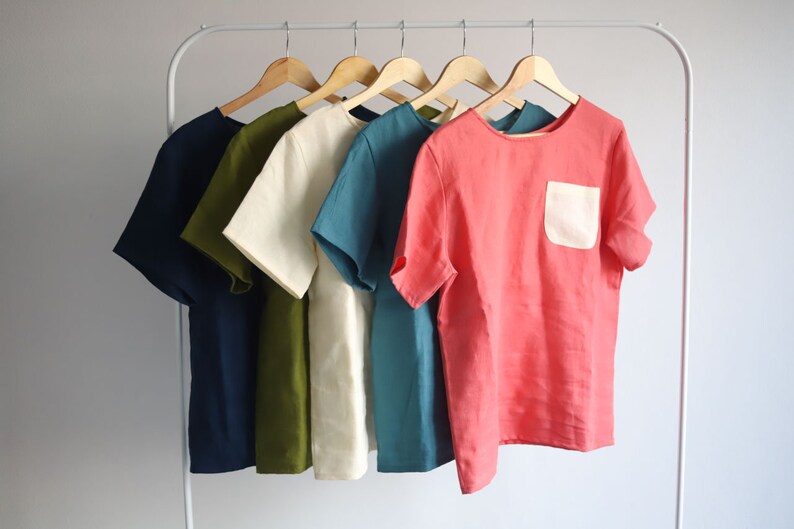 Pu&ograve; includere: Cinque camicie a maniche corte appese a grucce. Le camicie sono di vari colori, tra cui blu navy, verde oliva, crema, blu verde e corallo. Una camicia corallo ha una tasca a toppa bianca.