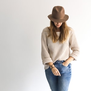 May include: A person wearing a brown fedora hat, a beige waffle-knit sweater, and blue jeans. The hat is positioned low, partially obscuring the face. The sweater has long sleeves, and the jeans are a light wash.