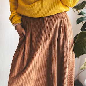 May include: A person wearing a mustard yellow knit sweater and a brown corduroy skirt. The skirt has vertical lines and a pleated design. The person has their hand in the skirt pocket. A plant is visible in the background.