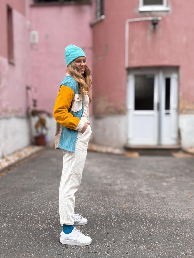 May include: A woman wearing a blue, yellow, and white corduroy jacket, white pants, and a blue beanie stands in front of a pink building. She is smiling and looking at the camera.