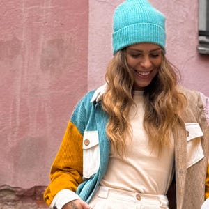 May include: A woman wearing a blue, white, and yellow corduroy jacket with a teal beanie. She is smiling and looking down.