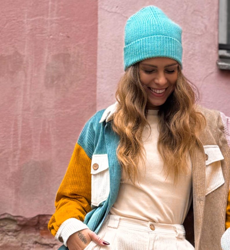 May include: A woman wearing a blue, white, and yellow corduroy jacket with a teal beanie. She is smiling and looking down.