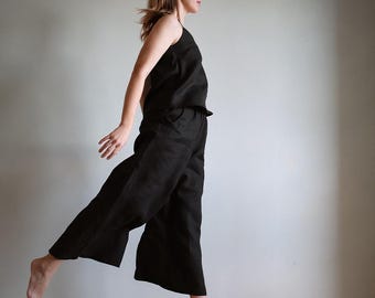 May include: Black linen camisole top and wide-leg pants. The outfit features a sleeveless top with thin straps and a high-waisted, loose-fitting pant. The model is captured mid-air, showcasing the flowy fabric and relaxed fit of the ensemble. The outfit is suitable for casual wear.
