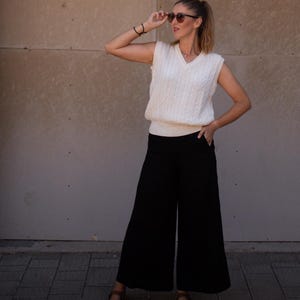 Puede incluir: Una mujer con un chaleco de punto trenzado color crema, pantalones negros de pernera ancha y sandalias marrones. Lleva gafas de sol y levanta la mano. El conjunto es sencillo y elegante.