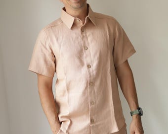 May include: A light peach linen shirt and shorts co-ord. The short-sleeved shirt has button fastenings and a collared neckline. The shorts have an elasticated waistband. The person is wearing a beige cap and glasses.