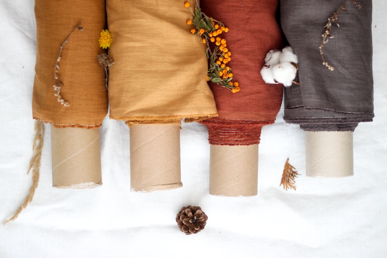 May include: Four rolls of fabric in different shades of brown, orange, and yellow, with dried flowers and cotton on top. The fabric is rolled up and sitting on a white surface.